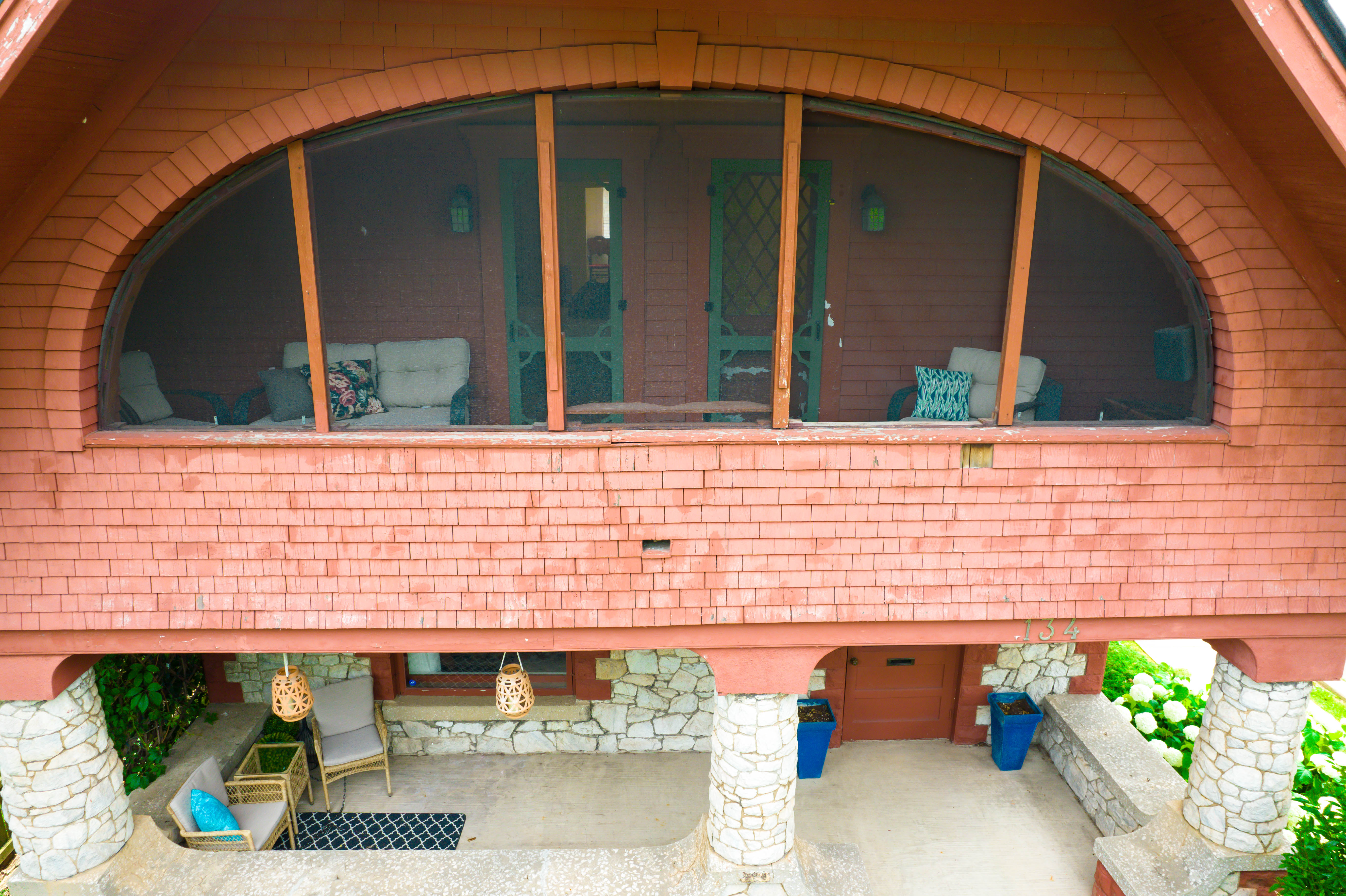 Exterior view of curved upper-level screened porch with stone pillars and red shake siding. Covered ground-level patio below adds extra outdoor space.