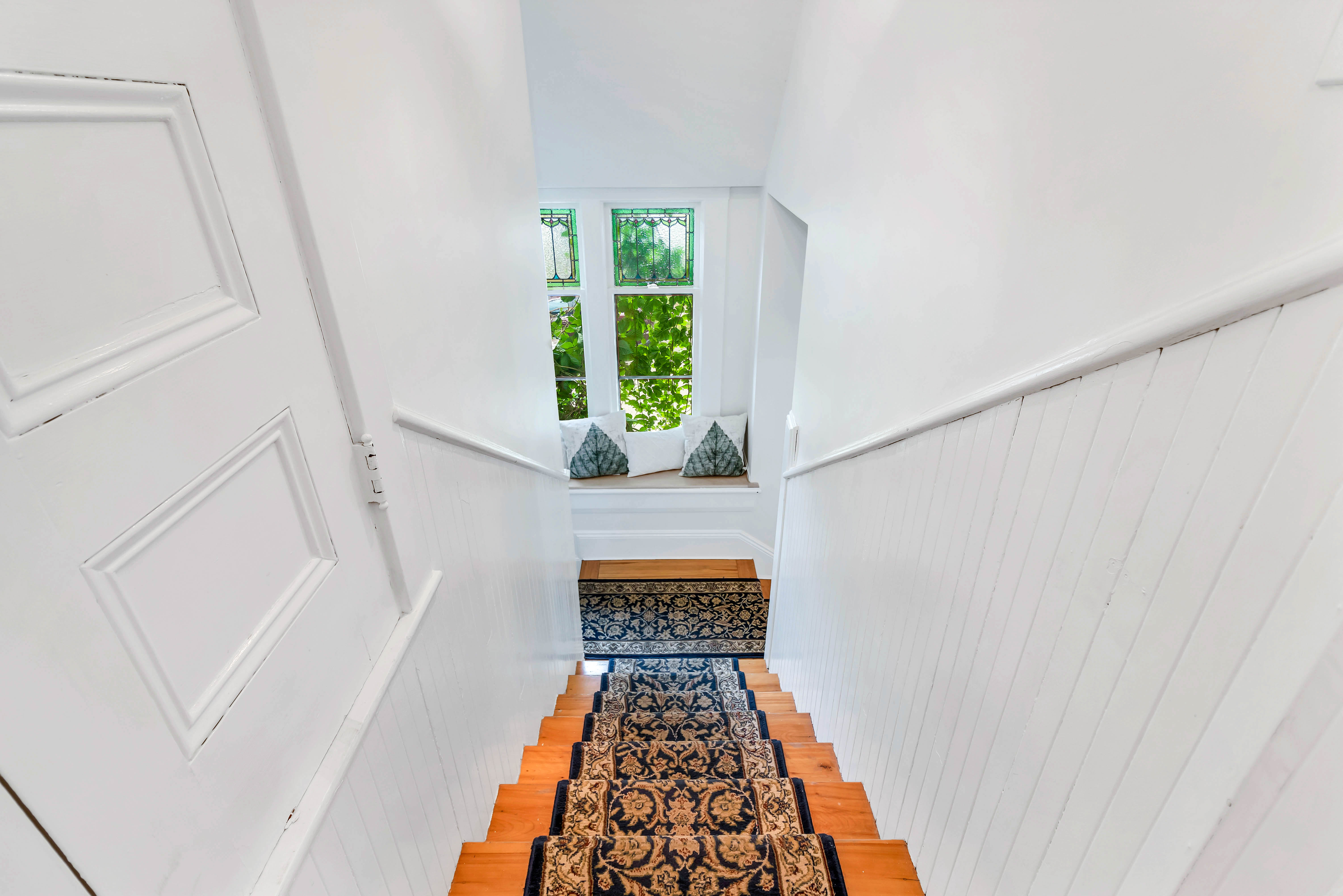 Top-down staircase view showcasing floral carpet runner, white wainscoting, and a large window seat at the landing with decorative stained glass.