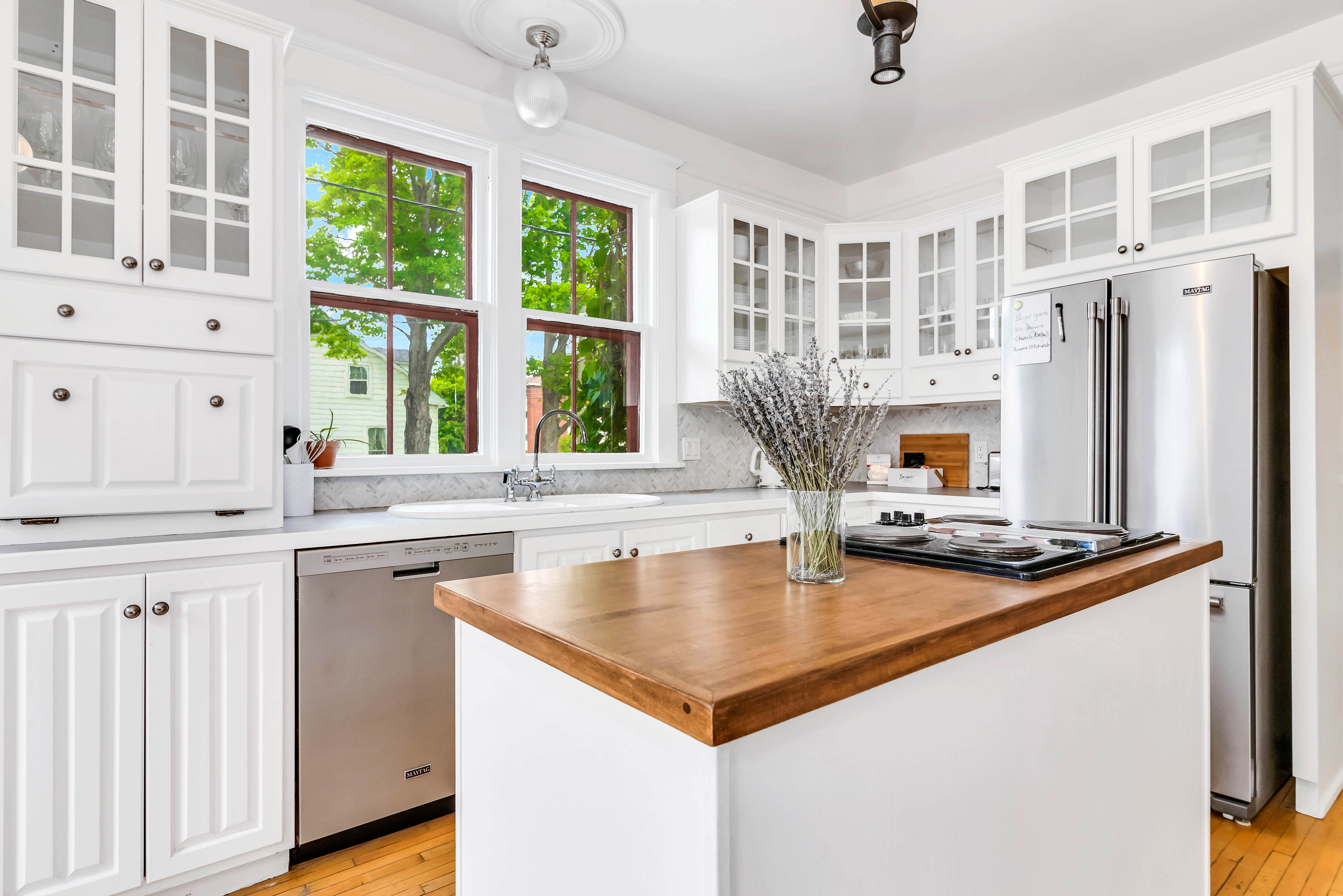 Sunny kitchen view with three large windows, white cabinetry with glass fronts, and stainless steel dishwasher. Center island holds a cooktop and vase of lavender.