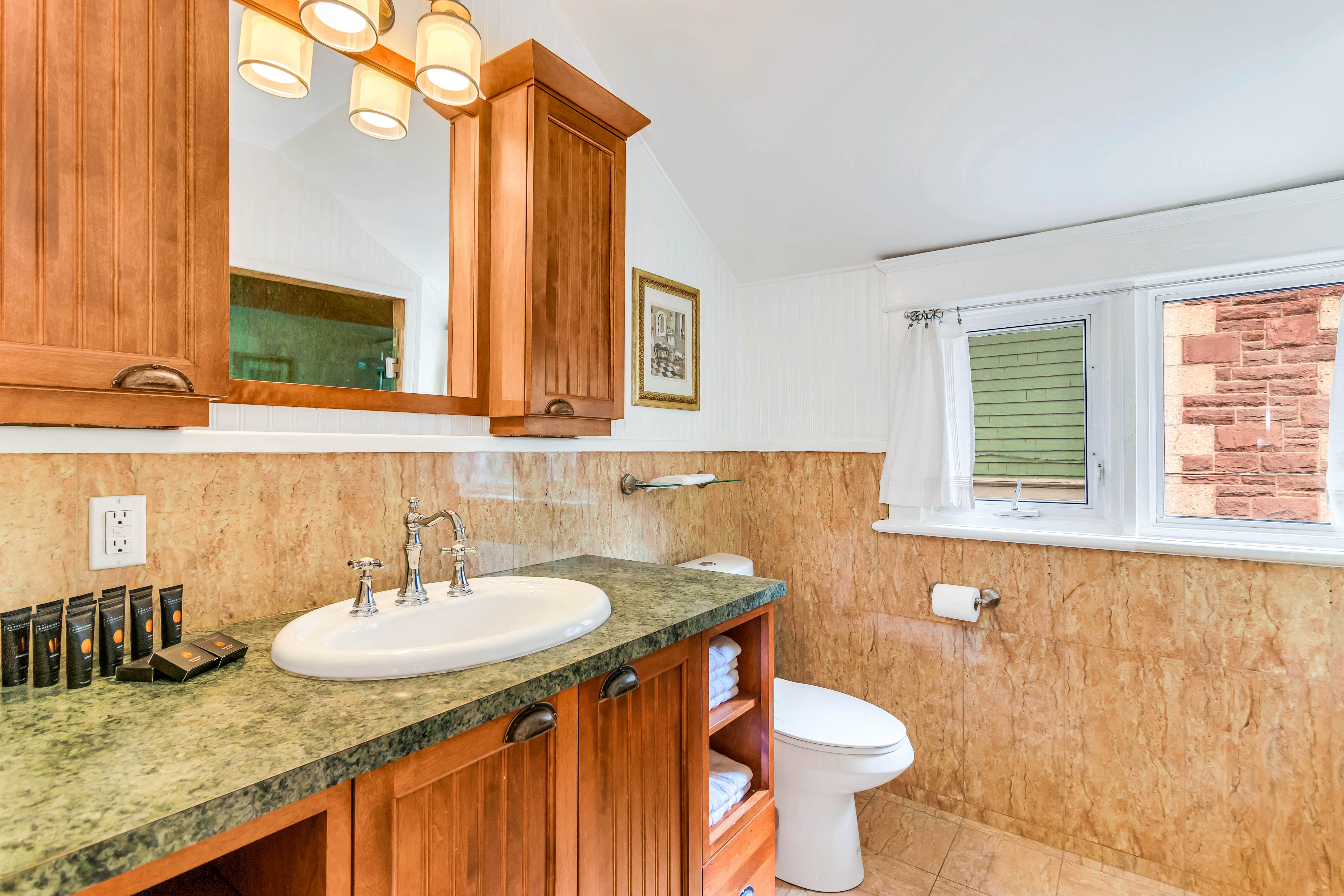 Charming bathroom with rich wood cabinetry, marble-look wall tiles, and a green stone countertop. Includes under-sink storage and natural light from dual windows.