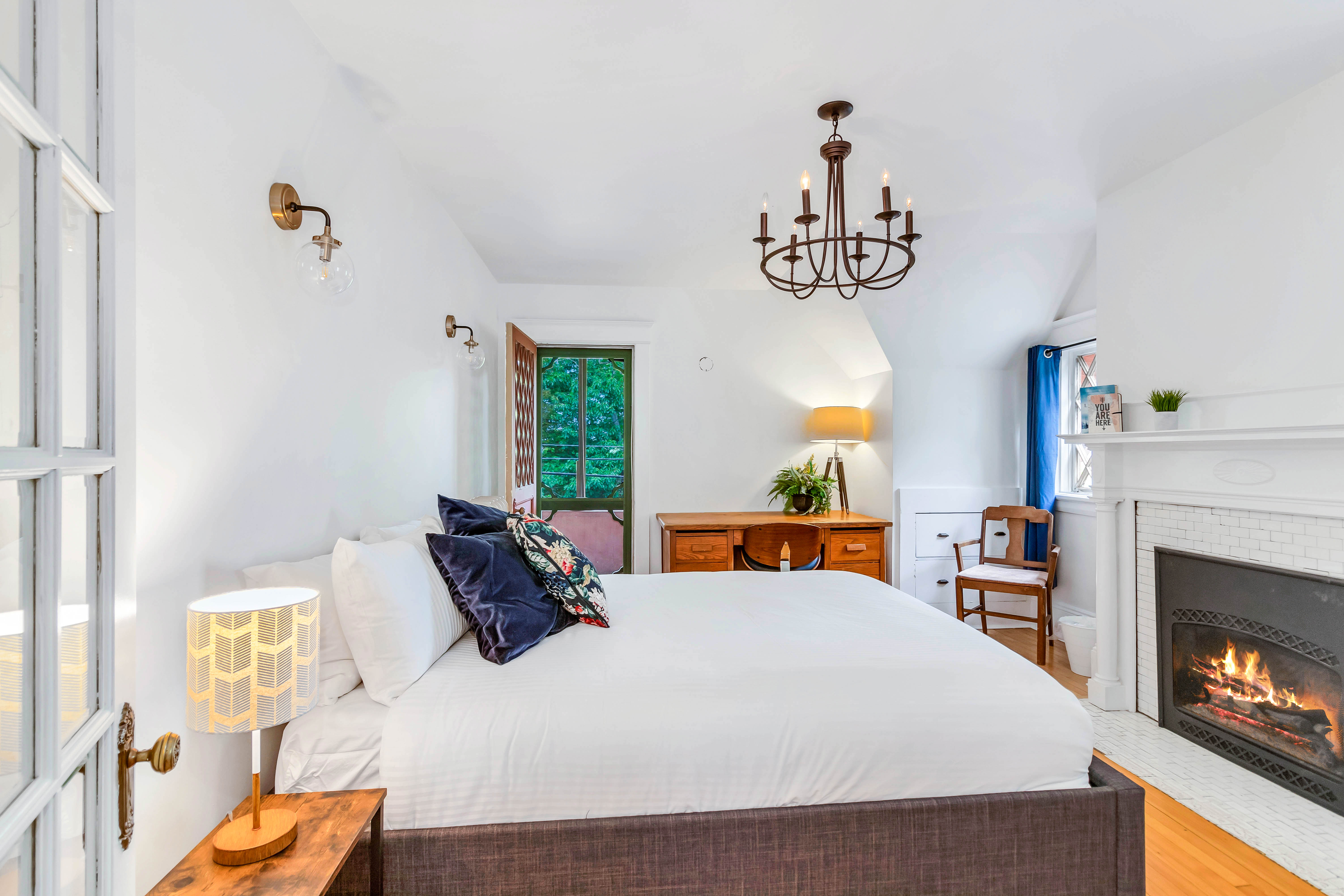 Elegant second bedroom with white walls, fireplace, floral throw pillows, and a wooden desk. French door opens to covered balcony.