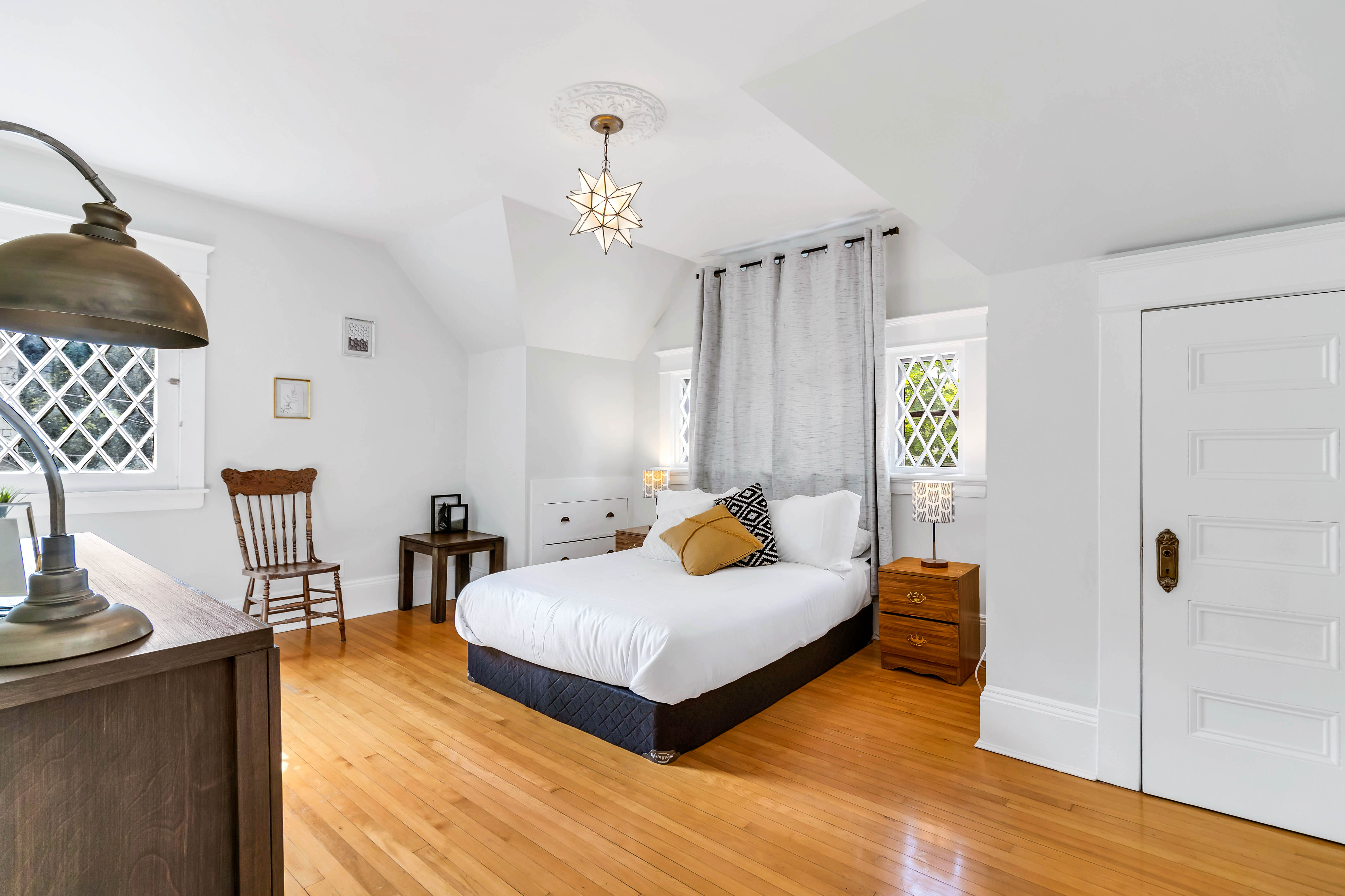 Guest bedroom with king-sized bed, vibrant throw pillows, neutral drapes, and a vintage red-upholstered wooden chair near a lattice window.