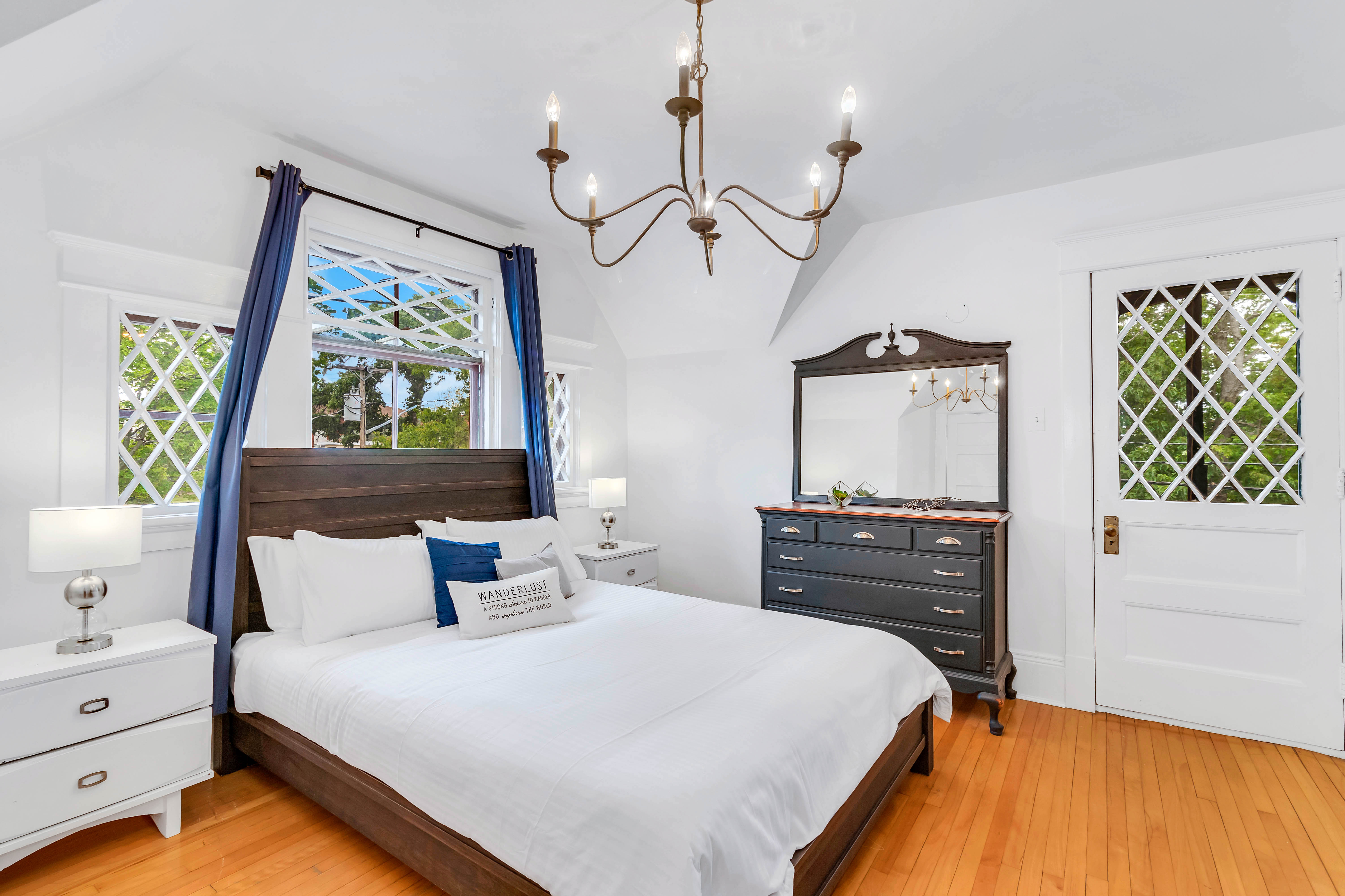 Primary bedroom with dark wood bed frame, white linens, vintage dresser, and lattice-style windows letting in natural light.