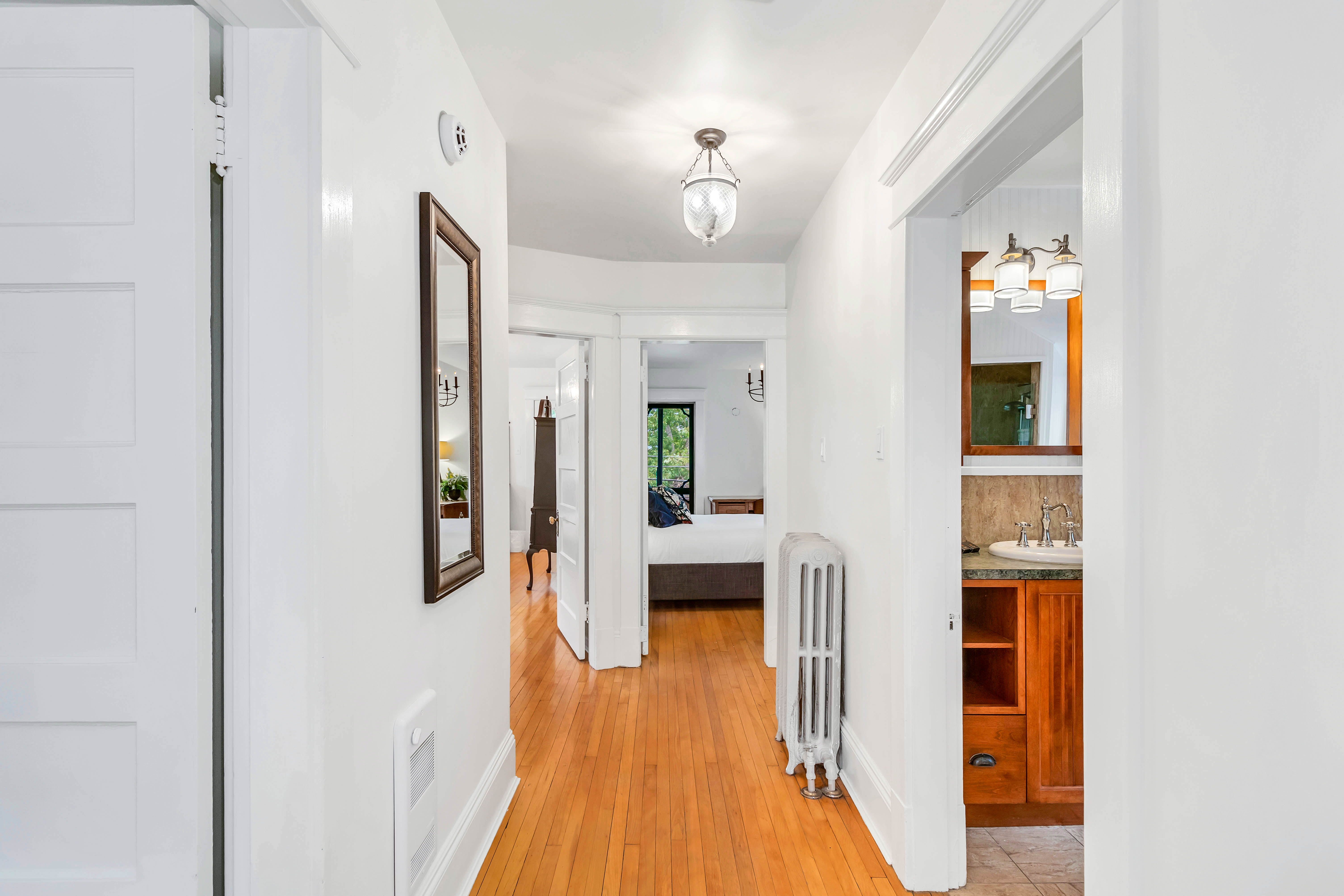 Upstairs hallway with original hardwood floors, leading to bedrooms and a bathroom. Features vintage radiator, wall mirror, and modern light fixture.