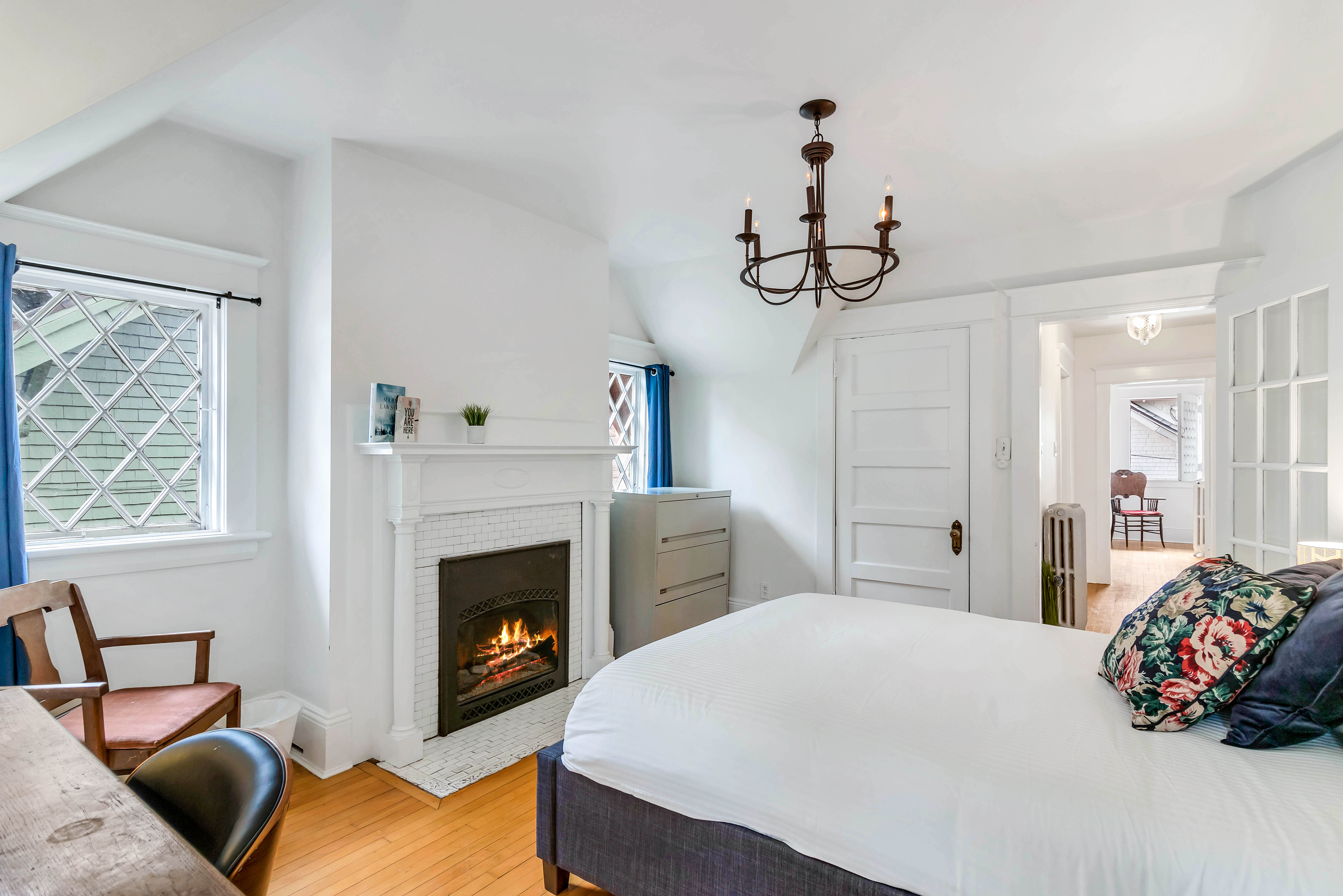Alternate angle of bedroom showing fireplace with white brick surround, blue curtains, and cozy seating area near the window.