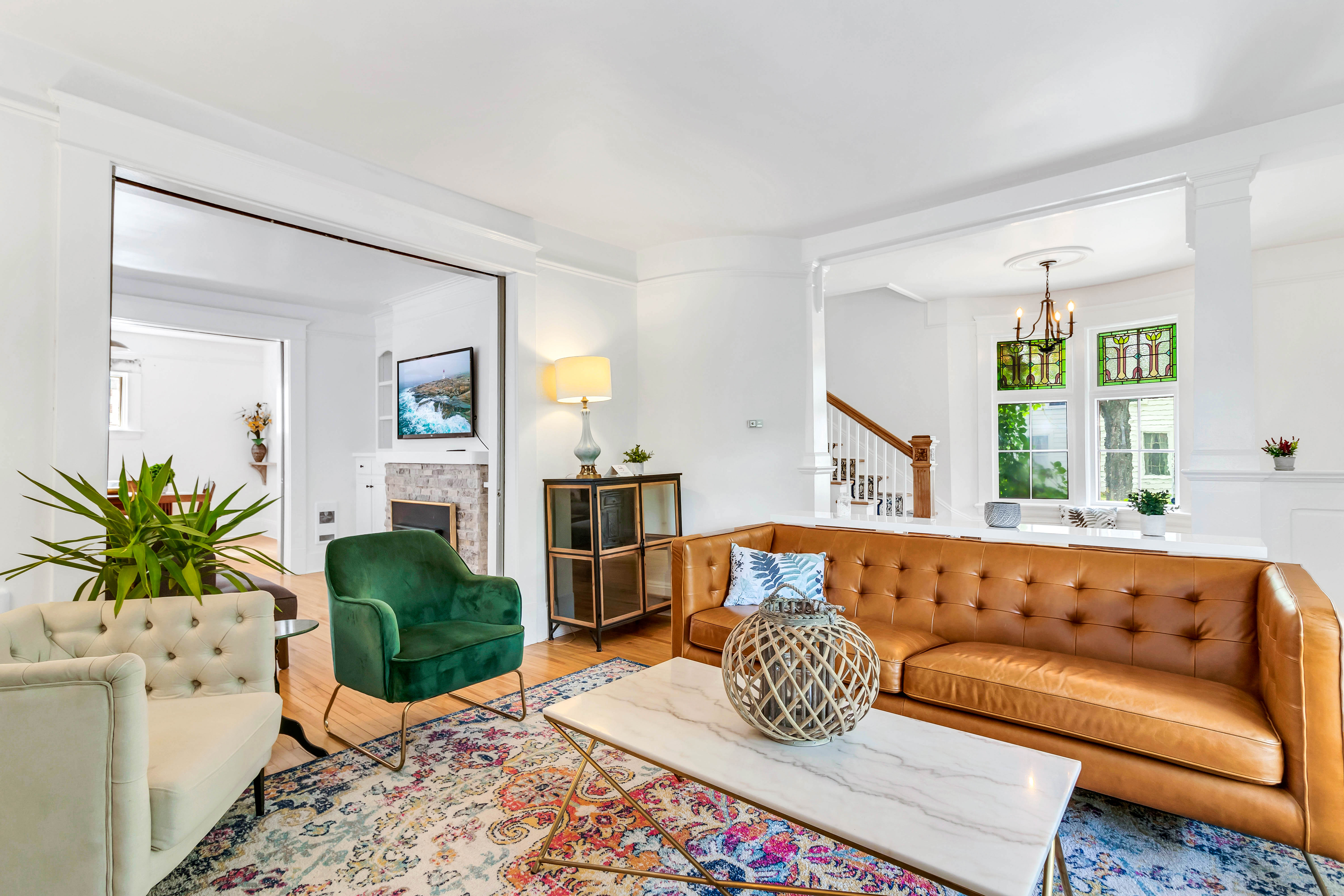 Cozy living room featuring a tufted leather sofa, velvet accent chairs, a colorful area rug, and large windows with stained glass.