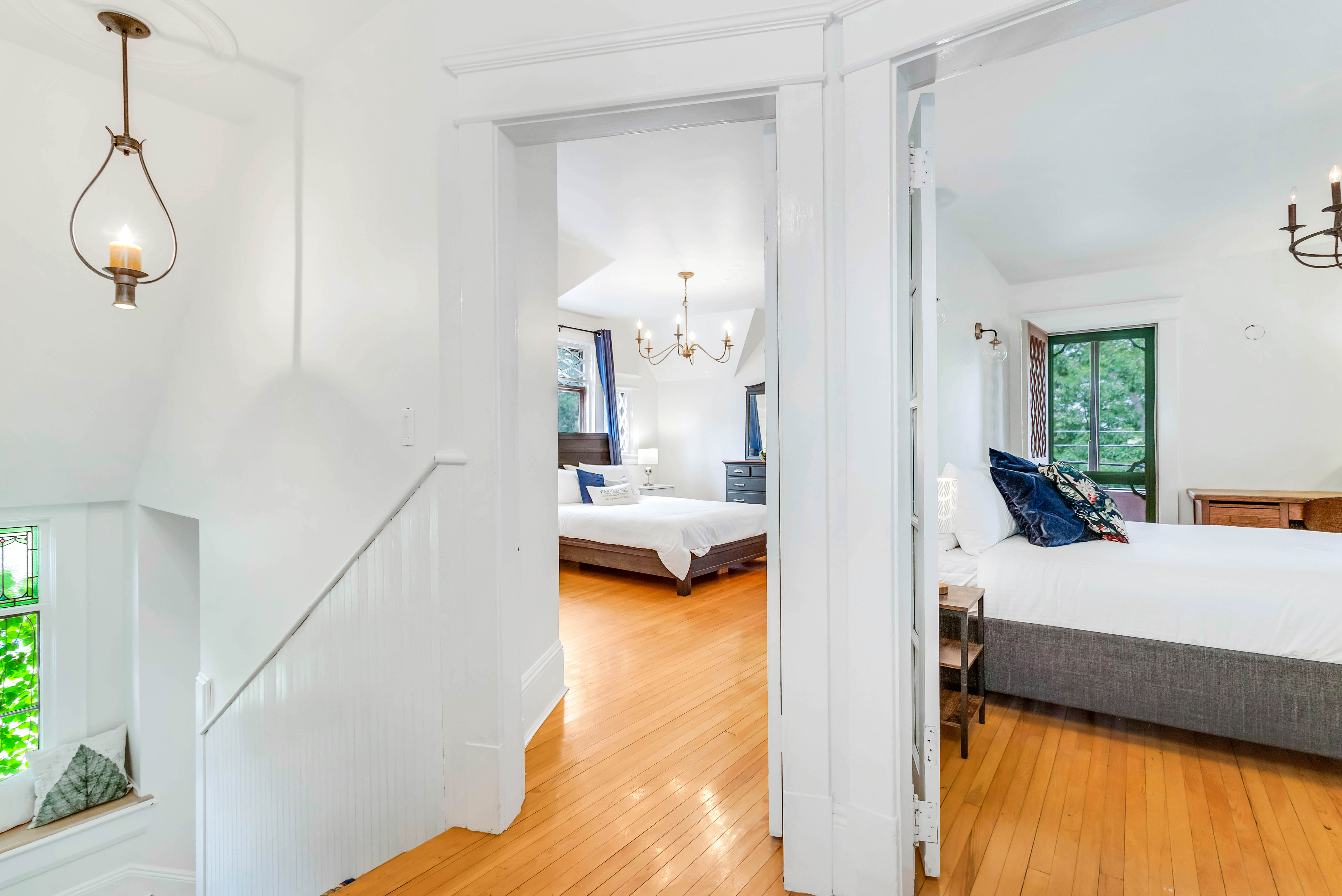 Second-floor hallway opening to two bedrooms, each featuring hardwood floors and elegant chandeliers. Stained glass window peeks from stairwell below.