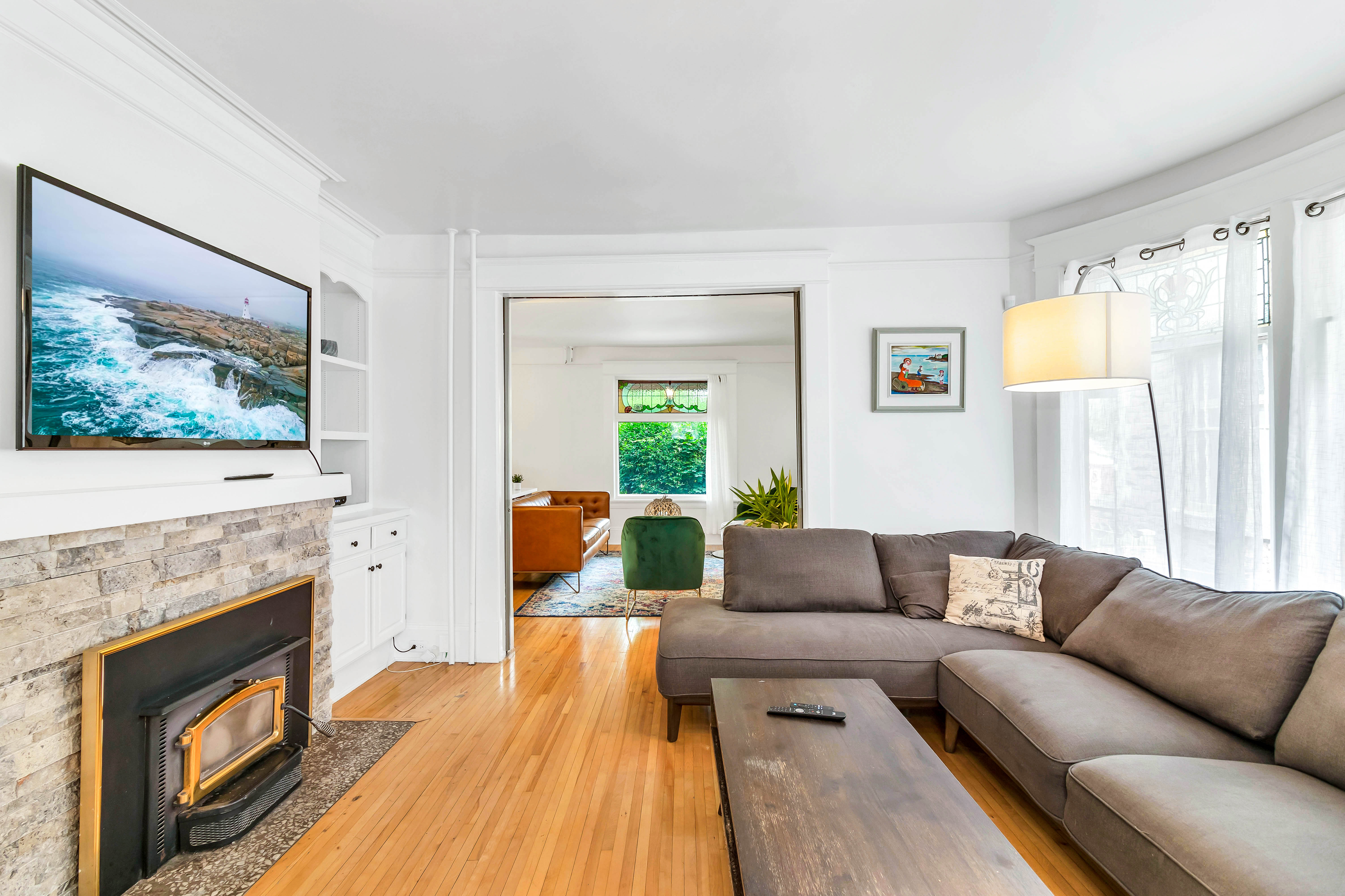 Secondary living room with grey sectional sofa, wall-mounted TV over a fireplace, and large curved windows with sheer curtains.