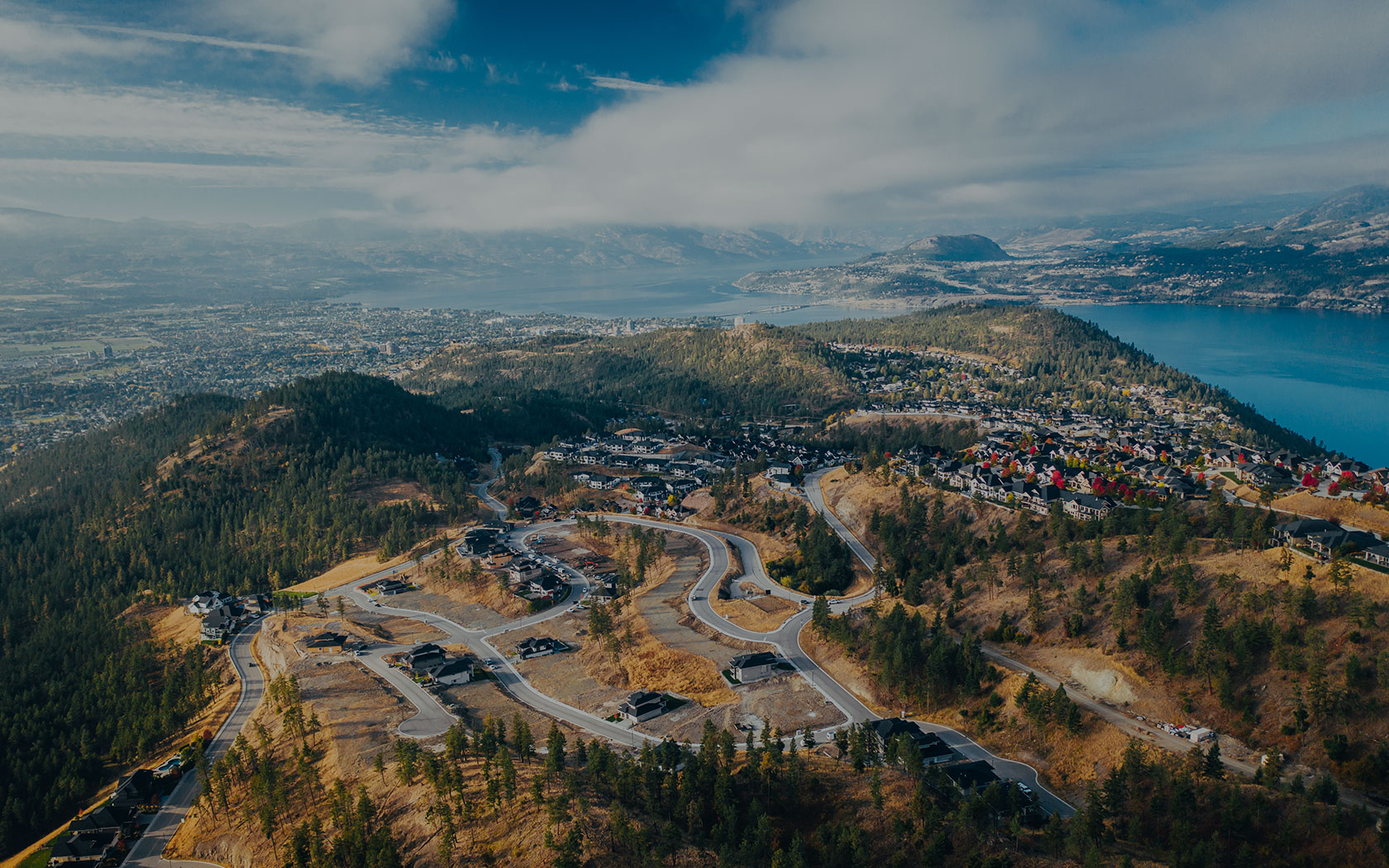 Aerial view of the Wilden master-planned community in Kelowna, showcasing luxury hillside homes and winding roads nestled in the Okanagan highlands. The photo highlights the neighborhood's natural parkland, energy-efficient architecture, and panoramic views of Okanagan Lake and downtown Kelowna in the distance under a blue sky.